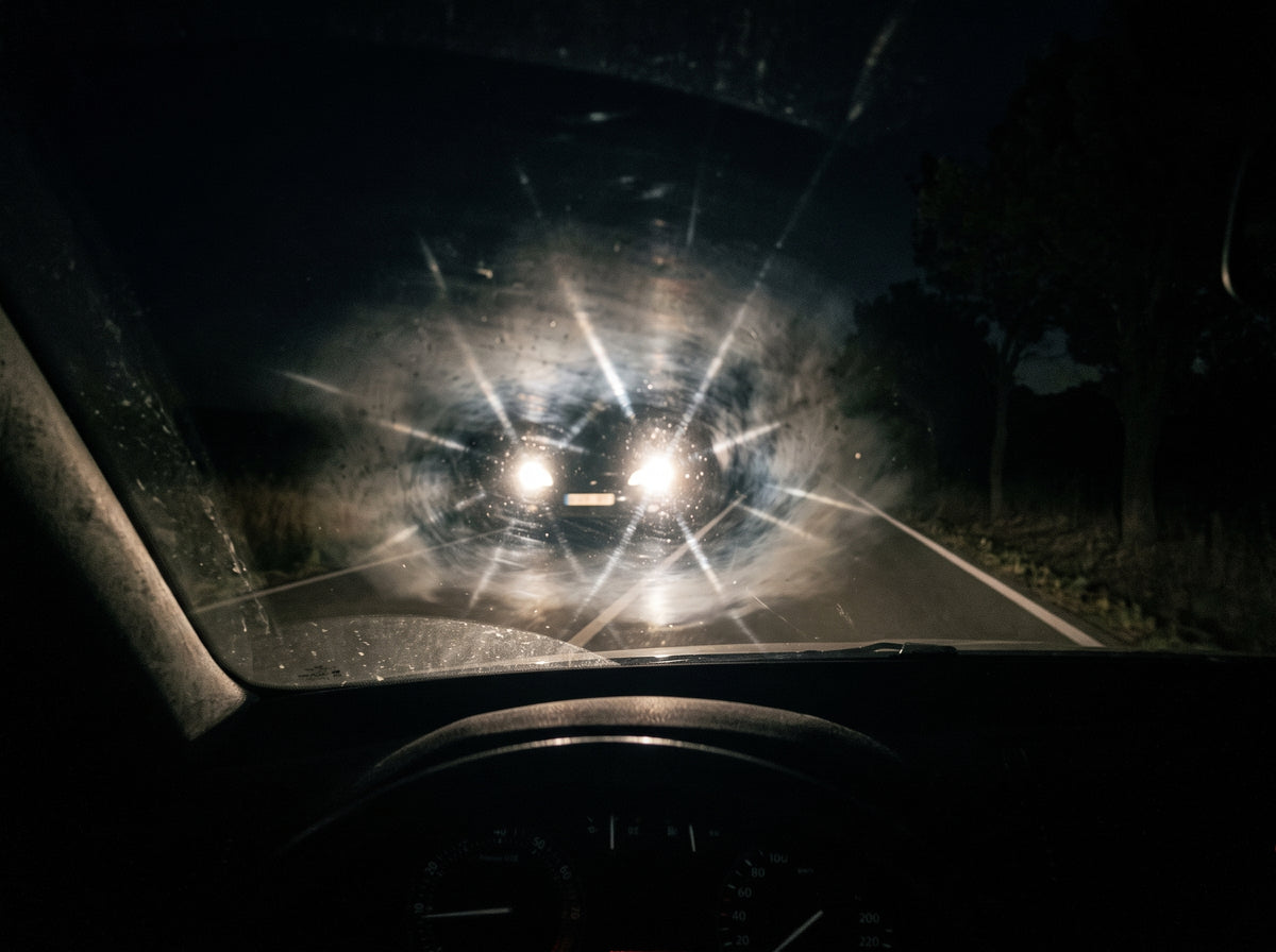 Faros de un coche de frente convertidos en halos a través de un parabrisas con grasa, de noche